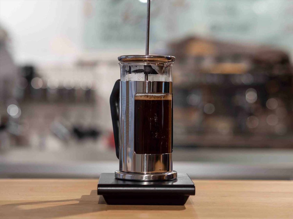 French Press aus Glas und Edelstahl mit Kaffee auf einer schwarzen Waage, Stempel noch nicht gedrückt